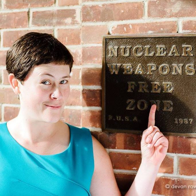 Brandi -- a white woman with dark hair and blue eyes -- points at a sign that states Nuclear Free Zone
